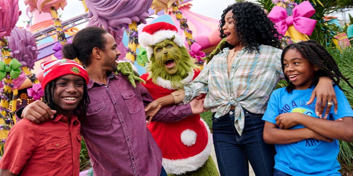 Four people smile and laugh as they pose with a person in a Grinch costume wearing a Santa suit, surrounded by colorful, whimsical decorations and large pink bows.