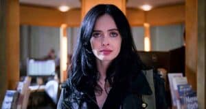 A woman with long black hair and a serious expression stands indoors, wearing a dark jacket. The background shows bookshelves and a softly lit room, suggesting a library or bookstore setting.
