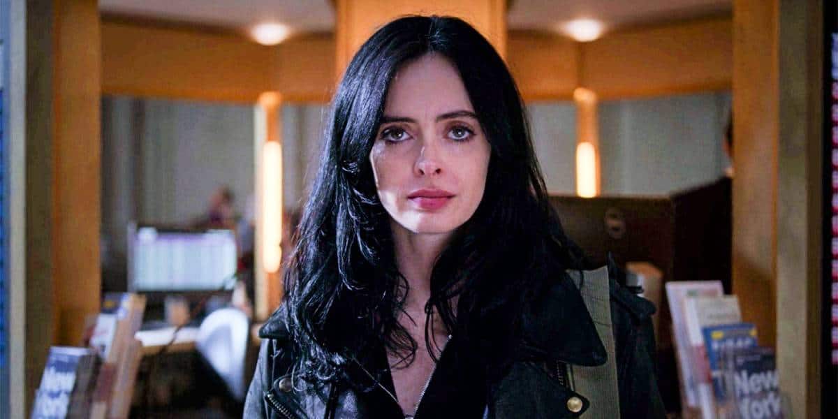 A woman with long black hair and a serious expression stands indoors, wearing a dark jacket. The background shows bookshelves and a softly lit room, suggesting a library or bookstore setting.
