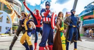 Six people in colorful Marvel superhero costumes, including Wolverine, Spider-Man, Captain America, Rogue, Storm, and Cyclops, pose heroically outdoors at a theme park with buildings and attractions visible in the background.