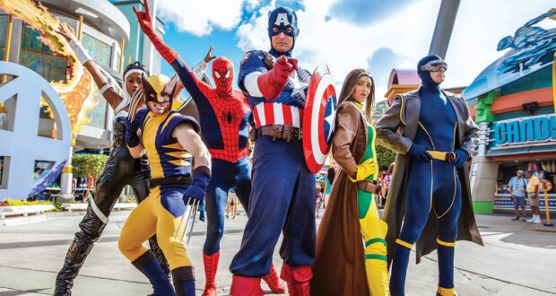 Six people in colorful Marvel superhero costumes, including Wolverine, Spider-Man, Captain America, Rogue, Storm, and Cyclops, pose heroically outdoors at a theme park with buildings and attractions visible in the background.
