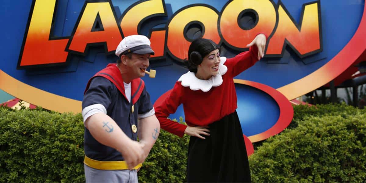 Two people dressed as Popeye and Olive Oyl pose in front of a colorful "LAGOON" sign, with greenery at their feet. Popeye wears a sailor outfit and pipe; Olive Oyl wears a red top with white collar and black skirt.