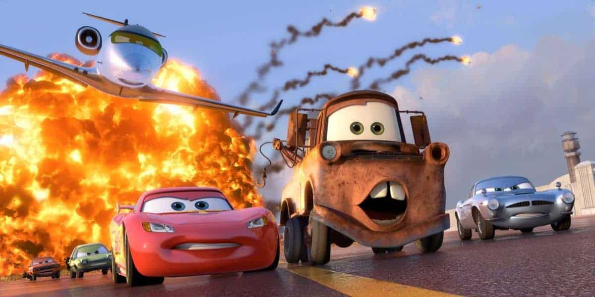 Animated cars with expressive faces speed down a road as a large explosion occurs behind them; a jet flies overhead with smoke trails. The scene is action-packed and colorful, set in the "Cars" movie universe.