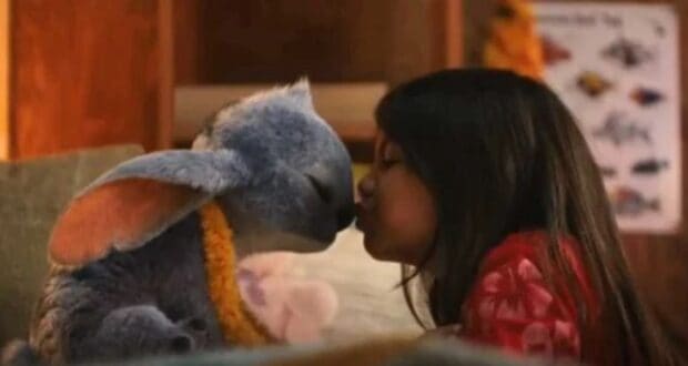 A young girl kisses a blue, furry creature with large ears and a lei around its neck. Indoors, they sit close in a warm setting—early reviews for Lilo and Stitch suggest $1 billion film magic for Disney.