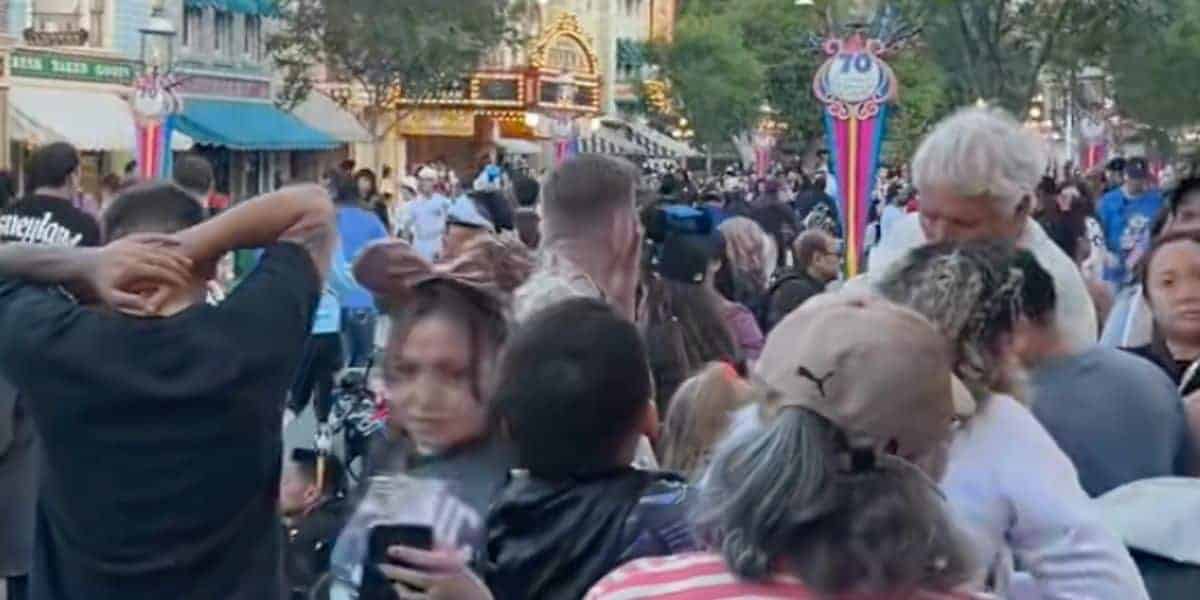 A crowded street scene at Disneyland, with people walking closely together, colorful banners, and illuminated buildings in the background. Trees line the street and park decorations are visible.