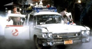 Two people in Ghostbusters uniforms step out of a white classic car with the Ghostbusters logo, surrounded by mist at night, with emergency lights and ghost-hunting equipment on the roof.