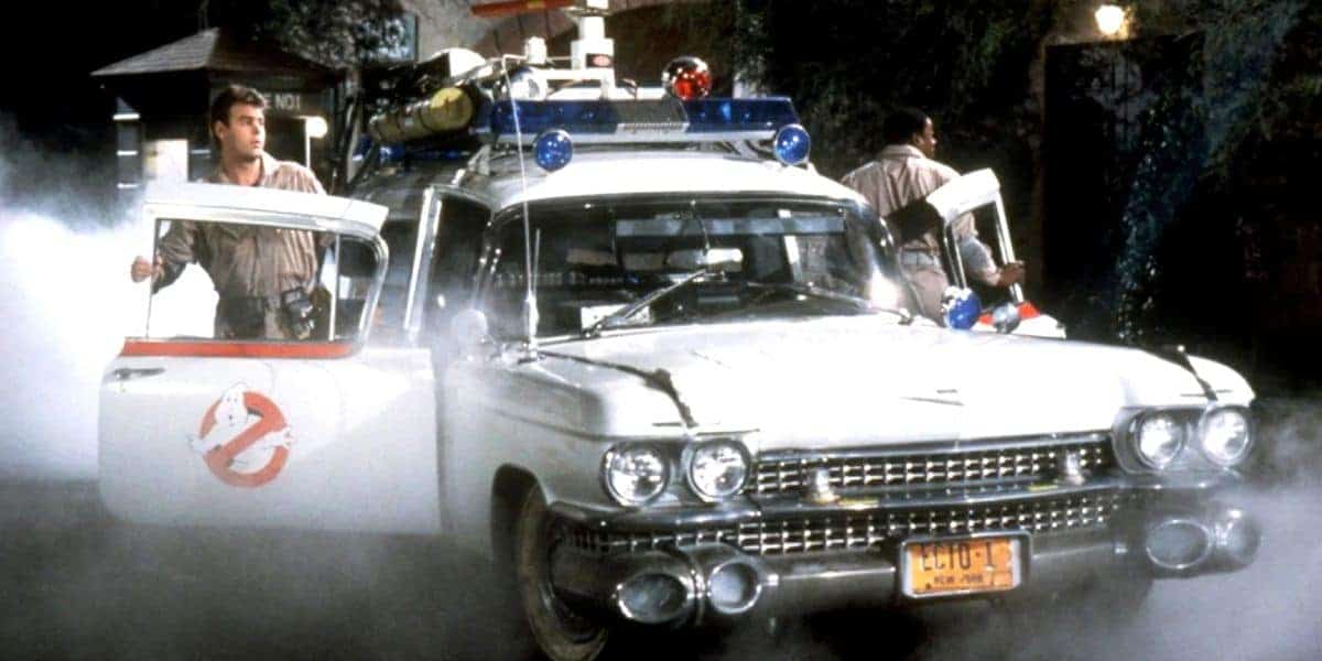 Two people in Ghostbusters uniforms step out of a white classic car with the Ghostbusters logo, surrounded by mist at night, with emergency lights and ghost-hunting equipment on the roof.