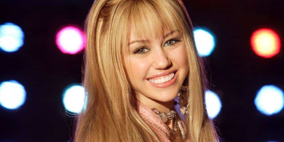 A young woman with long, straight blonde hair and bangs smiles at the camera. She is wearing a pink outfit, and colorful lights glow in the blurry background.
