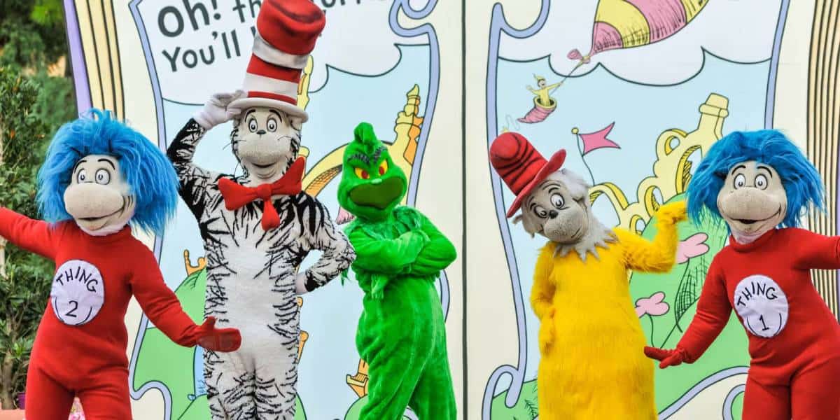 Four people in colorful Dr. Seuss character costumes, including Cat in the Hat, the Grinch, Thing 1, and Thing 2, pose in front of a large illustrated storybook backdrop outdoors.