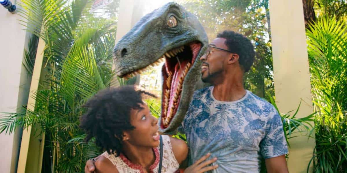 A couple stands in front of lush green plants, looking surprised as a large, realistic dinosaur model with an open mouth appears behind them.
