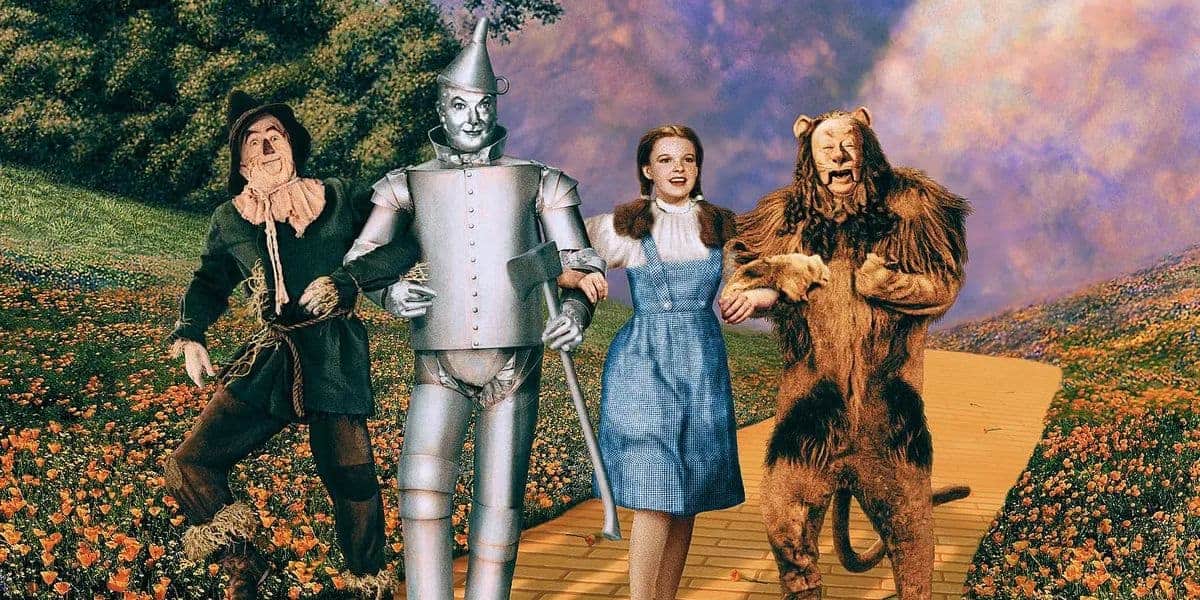Four characters—the Scarecrow, Tin Man, Dorothy, and Cowardly Lion—walk arm in arm down a yellow brick road, surrounded by colorful flowers and greenery, from "The Wizard of Oz.