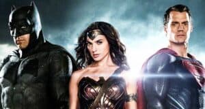 Batman, Wonder Woman, and Superman stand side by side in their superhero costumes against a dramatic, cloudy sky with light shining behind them.