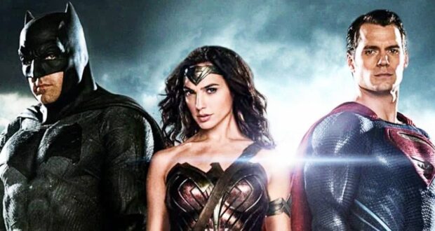 Batman, Wonder Woman, and Superman stand side by side in their superhero costumes against a dramatic, cloudy sky with light shining behind them.