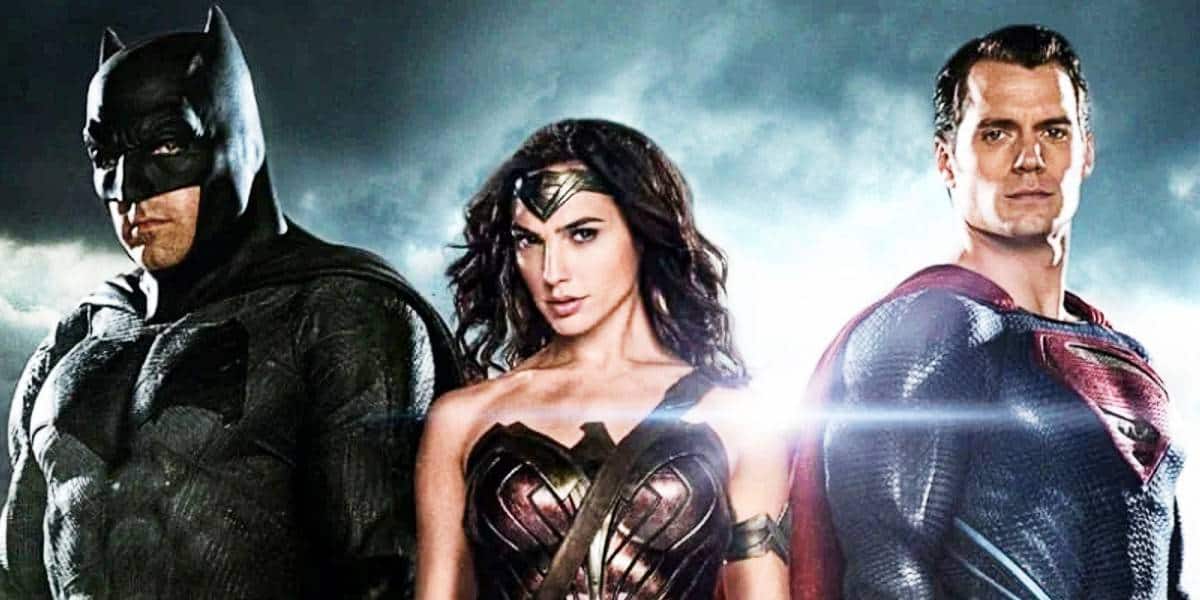 Batman, Wonder Woman, and Superman stand side by side in their superhero costumes against a dramatic, cloudy sky with light shining behind them.