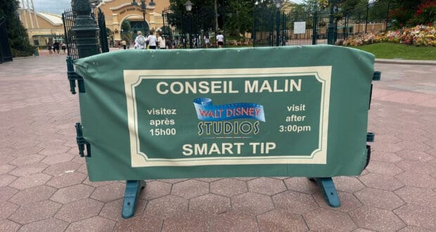 A green sign at the entrance to Walt Disney Studios Park reads "CONSEIL MALIN SMART TIP: visitez après 15h00 / visit after 3:00pm," advising visitors in both French and English.
