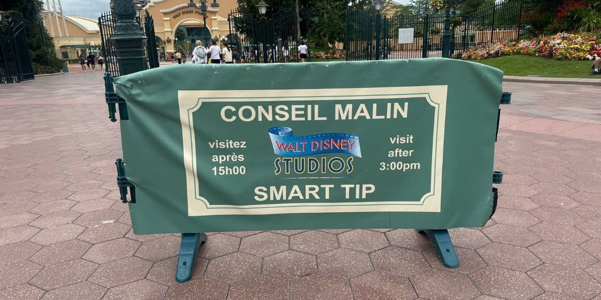 A green sign at the entrance to Walt Disney Studios Park reads "CONSEIL MALIN SMART TIP: visitez après 15h00 / visit after 3:00pm," advising visitors in both French and English.