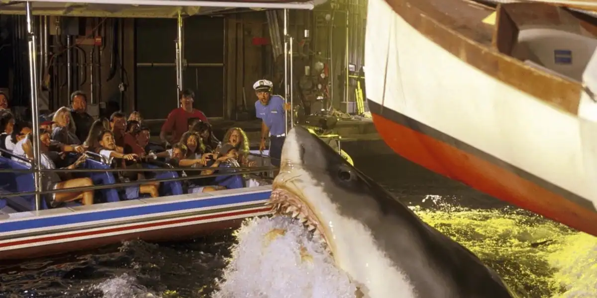 A mechanical shark emerges from the water near a boat, causing a group of tourists seated in a nearby tour boat to react with excitement and fear. The scene appears to be part of an amusement park attraction at Universal Orlando, with a staff member observing in the background.