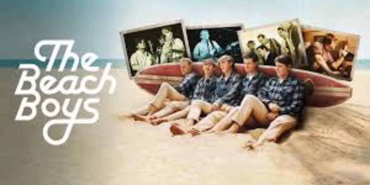 Five men in matching shirts sit on a surfboard in the sand, with "The Beach Boys" logo beside them. Behind them are photos of the band, including Brian Wilson, performing and together before Pet Sounds and their Disney movie collaborations.
