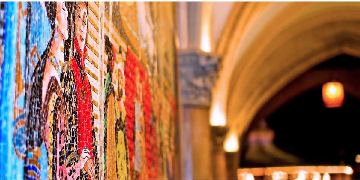 A colorful mosaic mural on a wall, featuring people in historic attire—echoing the style seen in the Nazi behind Disney World and Disneyland murals—with an arched hallway and warm lighting blurred in the background.