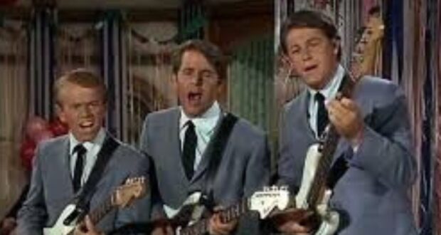 Three men wearing matching gray suits and black ties sing and play electric guitars together on stage, with colorful decorations in the background.