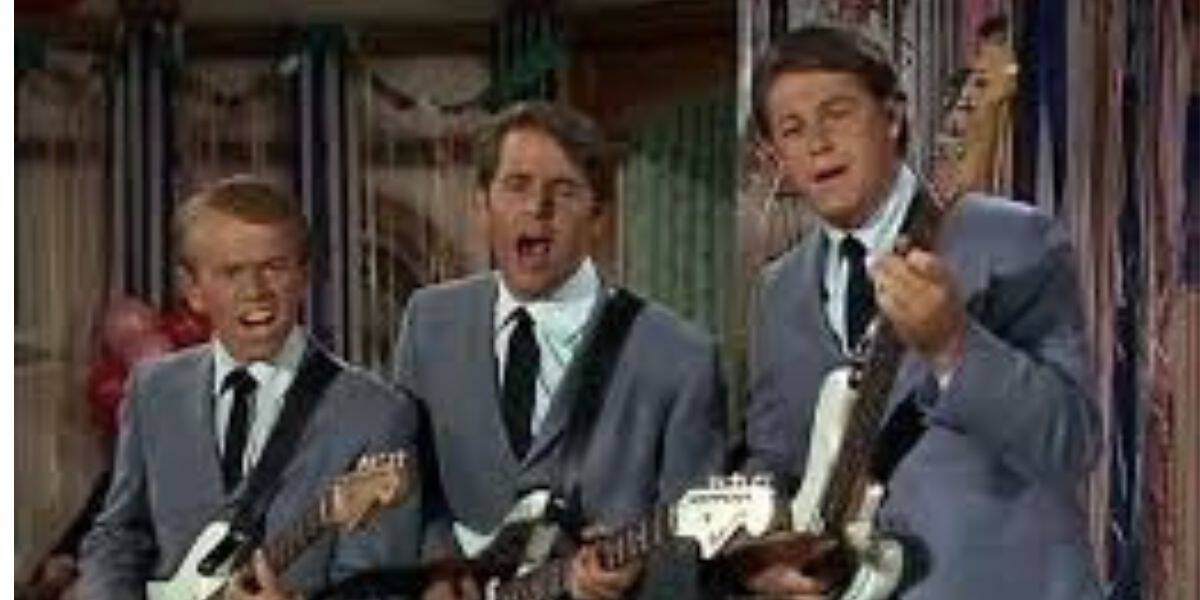 Three men wearing matching gray suits and black ties sing and play electric guitars together on stage, with colorful decorations in the background.
