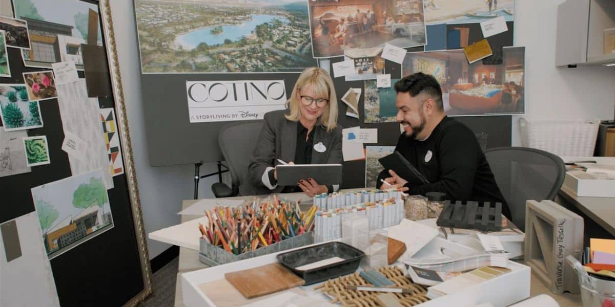 Two people sit at a desk covered with art supplies, sketches, and samples, discussing and smiling. Behind them are design boards and images—including a sign for "Cotino, a Storyliving by Disney"—and news that Disney North Carolina Asteria Community gets initial approval.