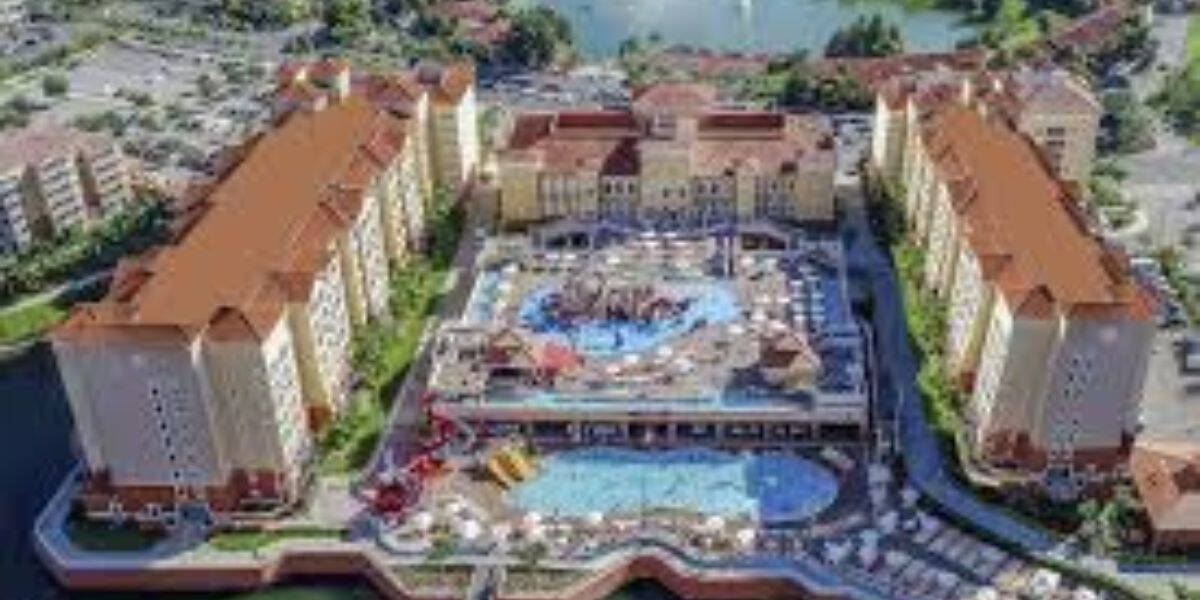 Aerial view of a large resort complex with two long, identical tan buildings facing a central courtyard with pools, lounge chairs, and umbrellas. Surrounding areas include parking lots, greenery, and a lake.