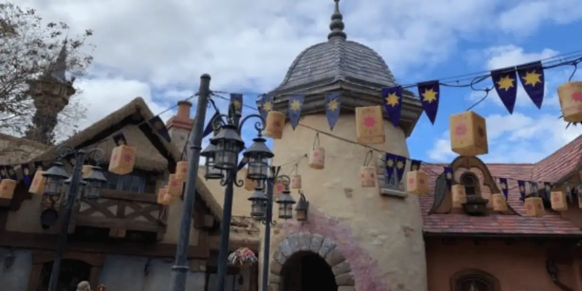 A whimsical building with a stone archway and turret, decorated with lanterns and purple banners featuring yellow sun symbols, set against a partly cloudy sky.
