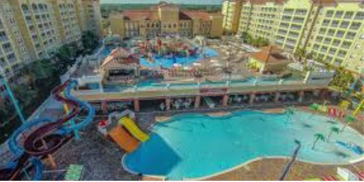 Aerial view of a large resort featuring multiple swimming pools, water slides, lounge chairs, and several tall yellow buildings surrounding the area on a sunny day.