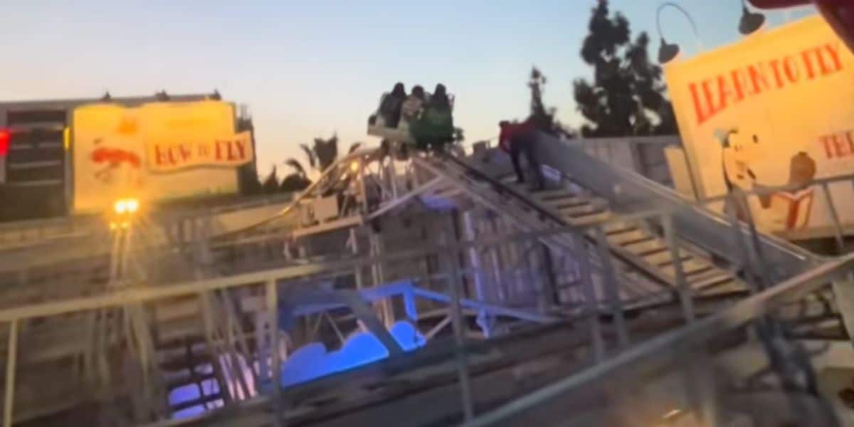 A group of people are stranded in a Goofy's Sky School car at the top of a track, as a person walks up the stairs to assist them