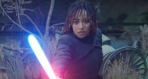 A determined woman with short, braided hair points a glowing blue lightsaber forward, standing amidst overgrown foliage and broken machinery in a dark, outdoor setting.