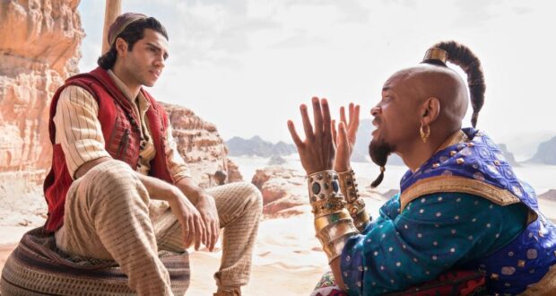 A man in striped pants and a red vest sits on a cushion in a desert landscape, listening intently to another man in ornate blue and gold clothing who gestures expressively with his hands.