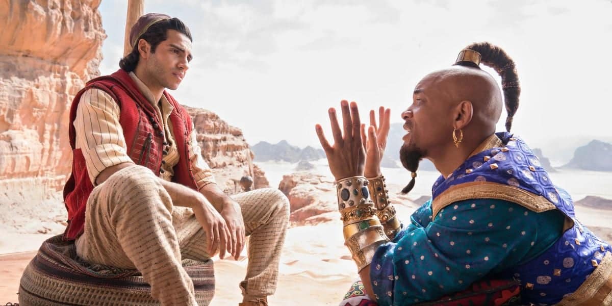A man in striped pants and a red vest sits on a cushion in a desert landscape, listening intently to another man in ornate blue and gold clothing who gestures expressively with his hands.