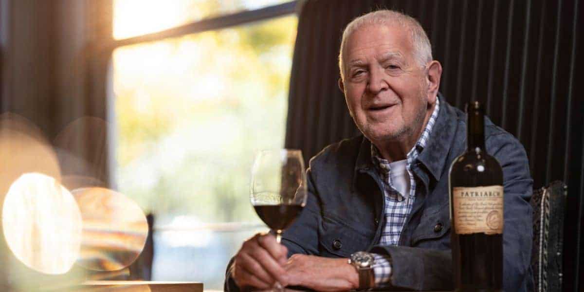 An older man sits at a table indoors, holding a glass of red wine and smiling. A bottle of wine is on the table next to him. Warm light comes through a window in the background.