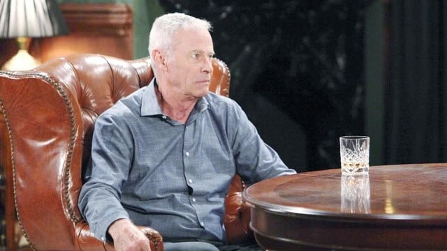 An older man with short gray hair sits in a brown leather armchair at a wooden table, wearing a blue shirt. A glass of whiskey is on the table in front of him. The background is dimly lit.