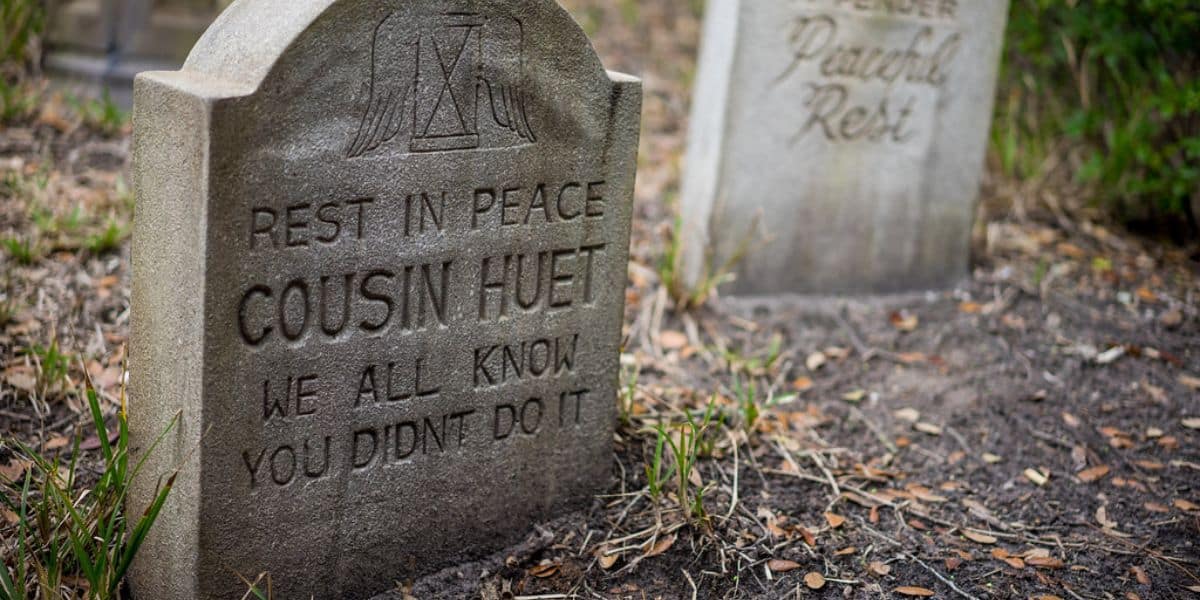 A gravestone reads, "Rest in peace Cousin Huet. We all know you didn't do it," with another blurred gravestone in the background and fallen leaves on the ground.
