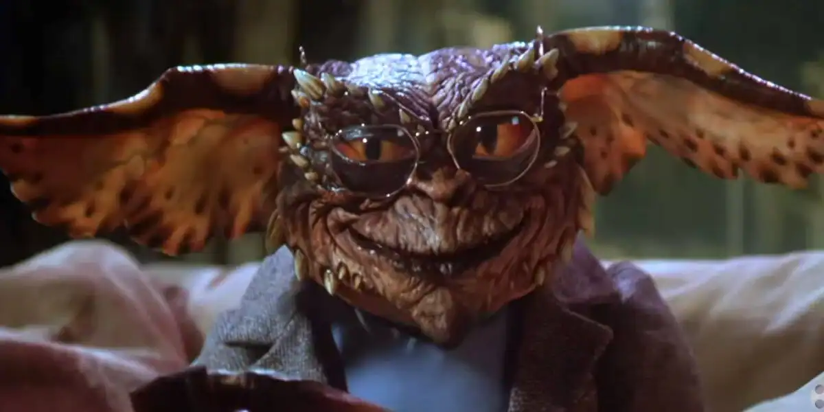 A gremlin creature with large ears, brown scaly skin, and glasses sits on a couch wearing a gray jacket, smirking at the camera.