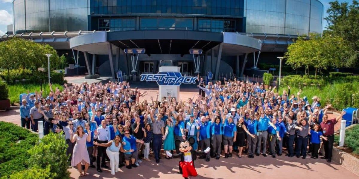 A large group of people, many wearing blue shirts, gather outside a building with "Test Track" signage. They are smiling, waving, and posing with a person dressed as Mickey Mouse in front. Lush greenery surrounds the area.