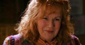 A woman with curly red hair smiles gently. She is wearing a colorful, patterned sweater and an orange scarf. The background is softly blurred, drawing focus to her warm expression.