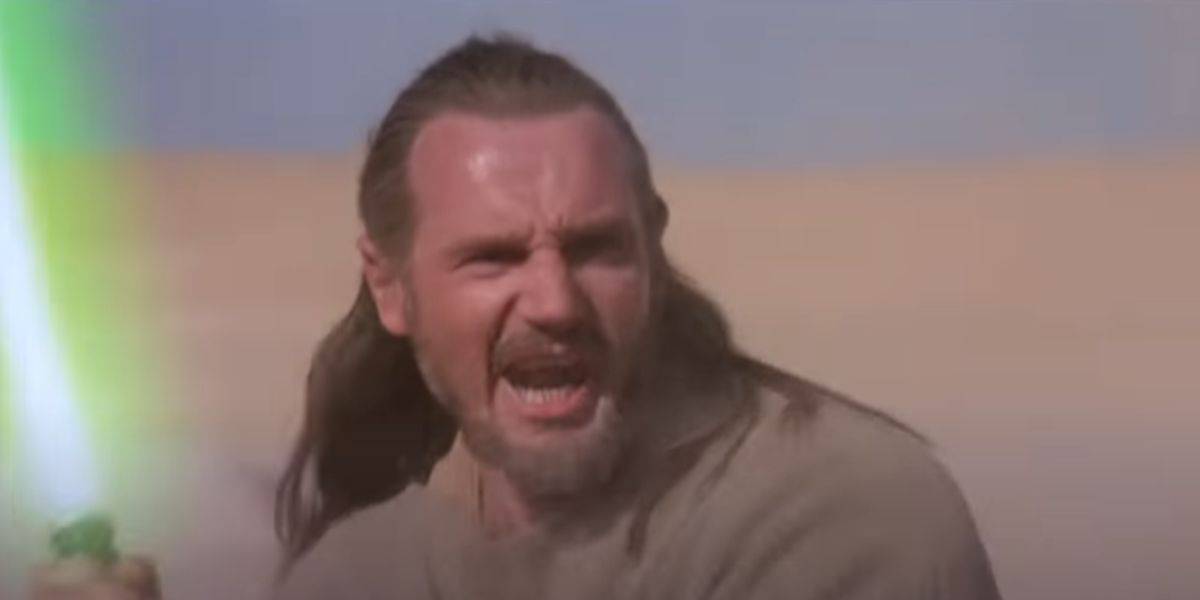 A bearded man with long hair shouts while holding a green lightsaber, standing in a desert landscape under a clear sky.