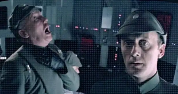 Two men in military uniforms stand in a dimly lit control room with glowing lights; one man is gasping and clutches his throat while the other looks forward with a serious expression.