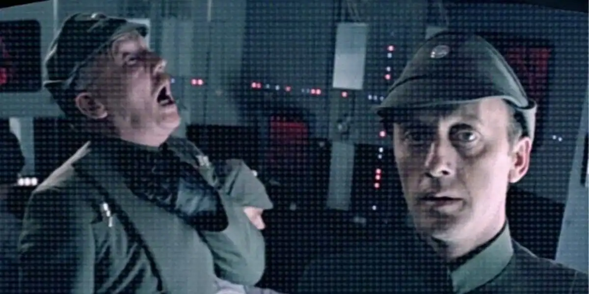 Two men in military uniforms stand in a dimly lit control room with glowing lights; one man is gasping and clutches his throat while the other looks forward with a serious expression.