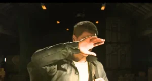A person in a dark jacket stands indoors, raising their hand to shield their face from a bright light, obscuring their facial features. The background is dimly lit, with industrial or warehouse elements visible.