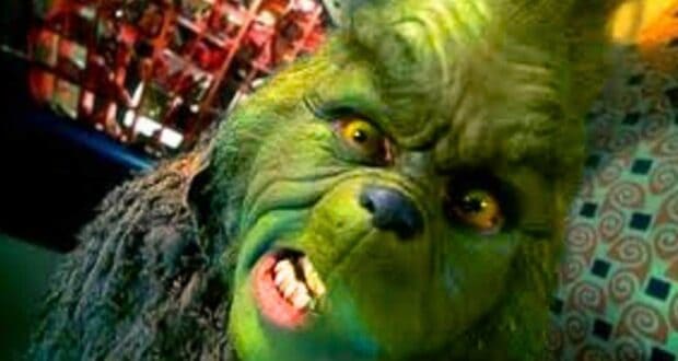 A close-up of a person in detailed green Grinch makeup and costume, baring their teeth and scowling intensely, with a colorful, blurred background.
