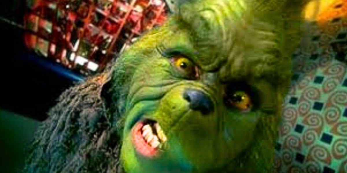A close-up of a person in detailed green Grinch makeup and costume, baring their teeth and scowling intensely, with a colorful, blurred background.