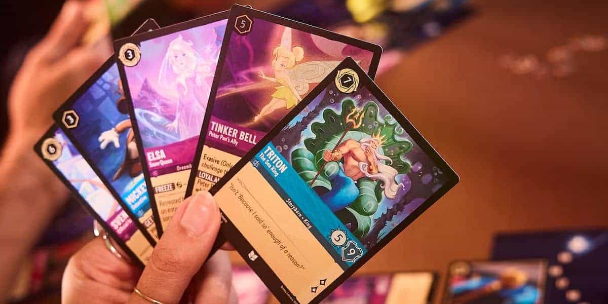A hand holding five colorful trading cards from a card game, featuring illustrated characters including Elsa, Tinker Bell, and others with fantasy-themed artwork and various game stats.
