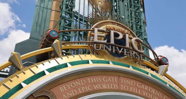 A close-up of the entrance sign for Universal Epic Universe, featuring gold, blue, and green decorative elements, with a blue sky and clouds in the background. Text below reads: "Beyond this gate find gardens and epic worlds to fill you.