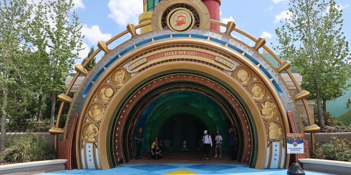 A colorful, round entrance tunnel at a theme park featuring Mario-themed decorations, gold accents, and cartoon character designs, with people entering and greenery in the background.