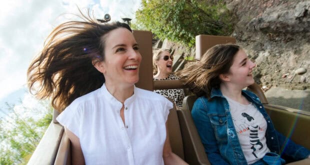 Three women ride a roller coaster outdoors, smiling and laughing as the wind blows their hair—a thrill much like Animal Kingdom attractions. The woman in front wears a white shirt, beside her is one in a denim jacket, and the third in back wears sunglasses.