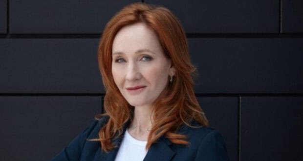 British author J.K. Rowling posing confidently, looking directly into the camera with red hair, a white blouse, and a black blazer against a black background. This image represents the Harry Potter author's confidence in an argument against Olympic medalist Imane Khelif, rekindling transphobic allegations against J.K. Rowling